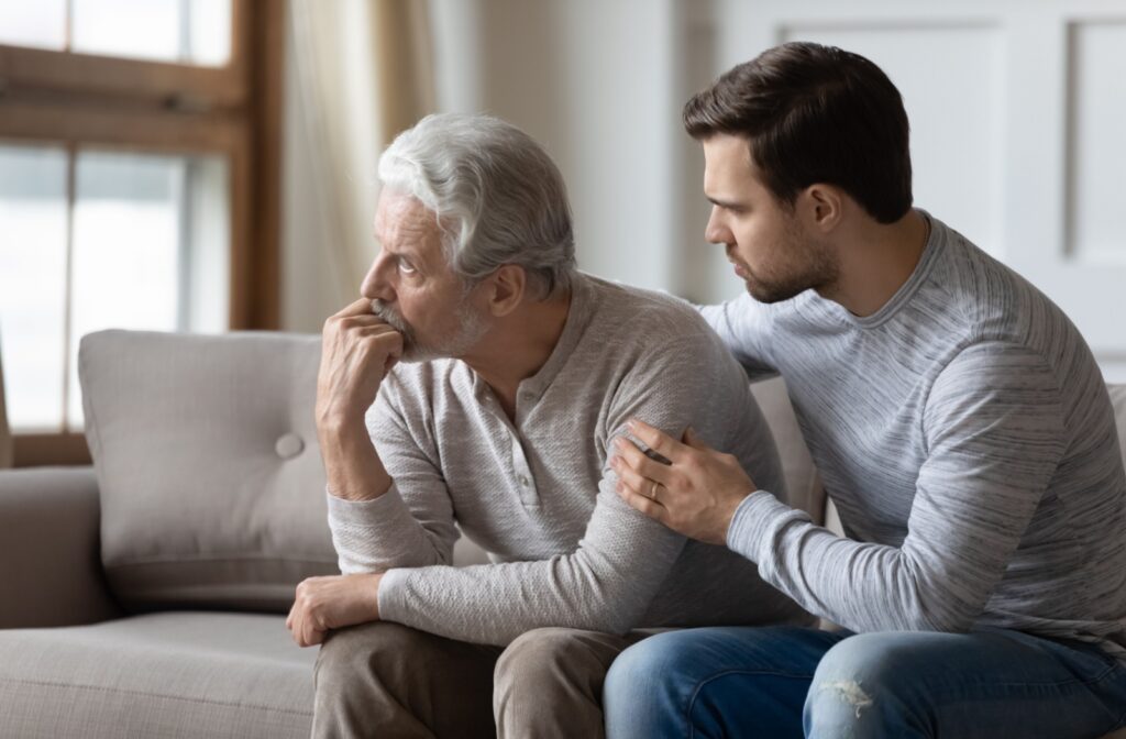 Concerned adult son comforting his senior father on a couch in a bright living room, showing support and discussing memory care needs