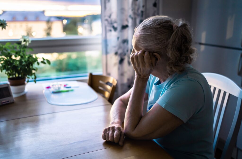 An older adult sits at their table and sadly looks out the window.