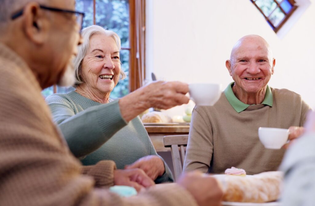 A group of seniors in the living room of a senior living community have tea and chat together while smiling and lifting their cups for a toast.