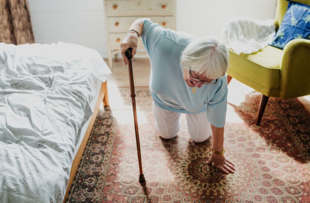 An older adult kneels on the rug in their bedroom and uses a cane to stand up after a minor fall